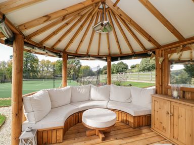 A wooden gazebo with a curved white cushioned bench and a round cushioned table overlooking a tennis court at Rockstowes House in Uley near Dursley
