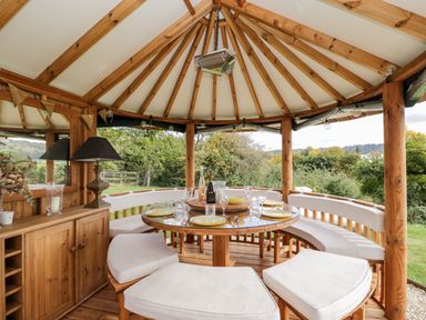 An outdoor wooden gazebo with a round dining table and cushioned curved bench seats at Rockstowes House Uley near Dursley