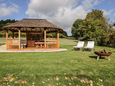 A wooden gazebo with seating on a lawn with two lounge chairs and a fire pit at Rockstowes House in Uley near Dursley