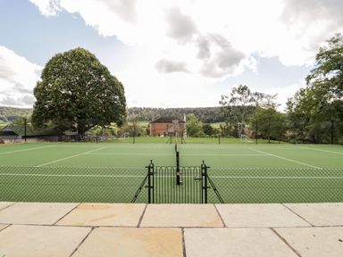 A tennis court with a black net and gate with trees and a house in the background at Rockstowes House in Uley near Dursley