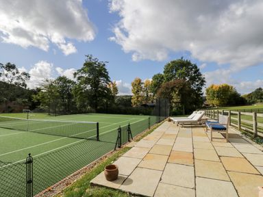 An outdoor tennis court next to a paved patio with lounge chairs and a bench at Rockstowes House Uley near Dursley