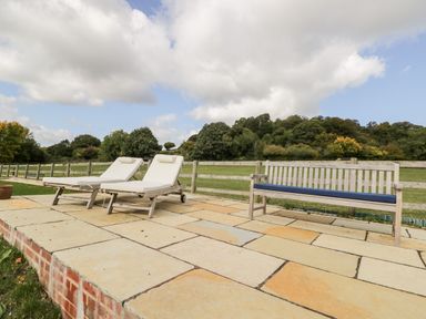 A patio area with two white cushioned sun loungers and a wooden bench with a blue cushion overlooking a fenced grassy field with trees at Rockstowes House Uley near Dursley