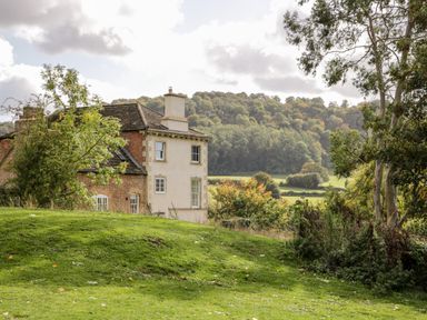 A countryside house with a grassy yard and trees with hills in the background at Rockstowes House in Uley near Dursley