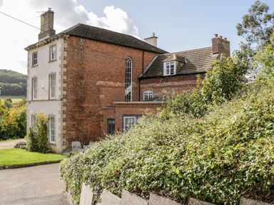 A brick and stone house with multiple windows and chimneys surrounded by bushes and trees at Rockstowes House in Uley near Dursley