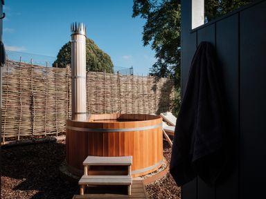 An outdoor wooden hot tub with steps and a tall chimney surrounded by a woven fence and trees at Rockstowes House in Uley near Dursley