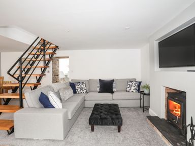 A living room with a grey sectional sofa and black ottoman near a fireplace and a staircase at Bodwena in Moelfre