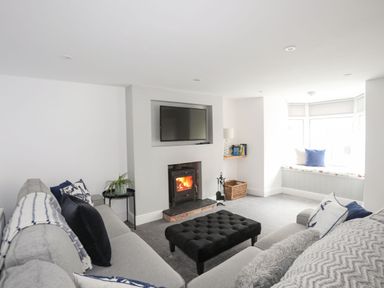 A living room with a wood burning stove a wall mounted tv a grey sofa a black ottoman and a window seat at Bodwena in Moelfre