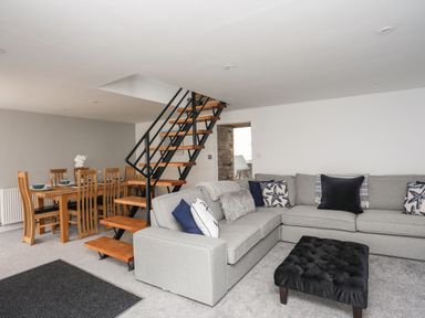 A living room with a gray sectional sofa and a black ottoman near wooden stairs and a dining table at Bodwena in Moelfre