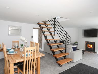 A dining area with a wooden table set with plates and glasses beside a staircase leading to an upper floor and a living area with a sofa a television and a fireplace at Bodwena in Moelfre