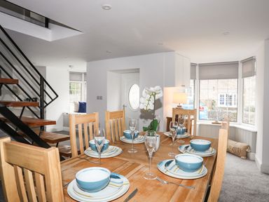 A dining area with wooden table and chairs set with plates bowls glasses and cutlery near staircase and window at Bodwena in Moelfre