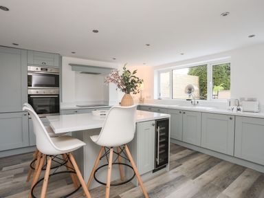A kitchen with light gray cabinets white countertops two white chairs and a large window at Bodwena in Moelfre