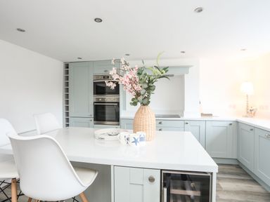 A modern kitchen with light blue cabinets white countertop a flower vase and two white chairs at Bodwena in Moelfre