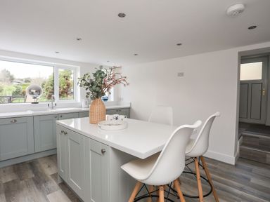 A kitchen with grey cabinets a white countertop a vase with flowers and three white chairs at Bodwena in Moelfre