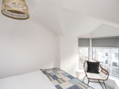 A bedroom corner with a bed, a patchwork blanket, a wicker chair with cushion by windows with grey blinds at Bodwena in Moelfre