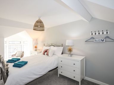 A bedroom with a white bed and teal towels on it two white dressers with lamps and a wicker chair near a window at Bodwena in Moelfre