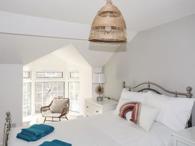 A bedroom with a metal bed frame white bedding and a wooden chair by a bay window at Bodwena in Moelfre