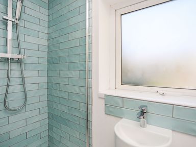 A shower area with blue-green wall tiles and a window above a sink with a chrome faucet at Bodwena in Moelfre