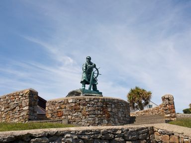 A statue of a man steering a ship's wheel on a stone platform with stairs and palm trees in the background at Bodwena in Moelfre