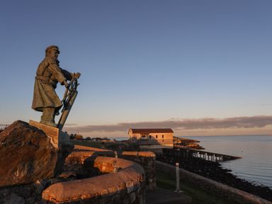 A statue of a man steering a ship wheel near a stone wall by the sea with a building and pier in the distance at Bodwena in Moelfre