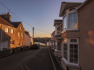 A street with houses on both sides and a car parked on the left side near the water at Bodwena in Moelfre