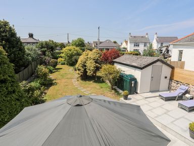 A garden with a stone path trees and a shed next to a patio with lounge chairs and an umbrella at Bodwena in Moelfre