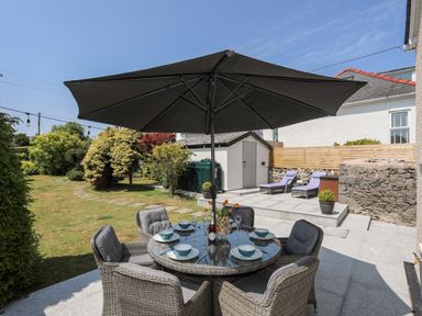 An outdoor patio with a round table set with dishes and glasses under a large umbrella next to sun loungers and a garden at Bodwena in Moelfre