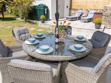 An outdoor patio dining area with a round glass table set with plates bowls glasses and a bottle surrounded by six cushioned wicker chairs at Bodwena in Moelfre