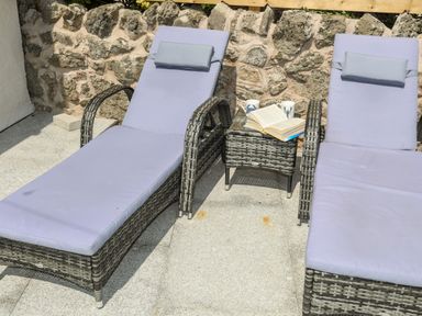 Two patio lounge chairs with cushions and a side table with books and mugs on a stone patio at Bodwena in Moelfre
