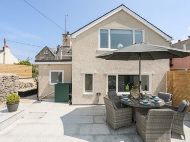 An outdoor patio area with a round glass table and six wicker chairs under a large umbrella at Bodwena in Moelfre
