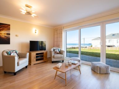 A living room with two armchairs a wooden TV stand a coffee table and large sliding glass doors overlooking a garden and the sea at Blue Ridge in Polzeath