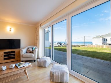 A living room with a chair a wooden TV stand a television and two poufs by large sliding glass doors with a view of a garden and houses at Blue Ridge in Polzeath