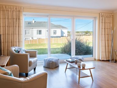 A living room with armchairs a coffee table with mugs and books large glass doors with curtains at Blue Ridge in Polzeath