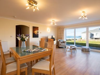 A dining area with wooden table and chairs next to a living room with armchairs and coffee table with sea view through sliding glass doors at Blue Ridge in Polzeath