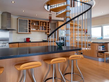 A kitchen with wooden cabinets black countertops a spiral staircase and three wooden bar stools at Blue Ridge Polzeath