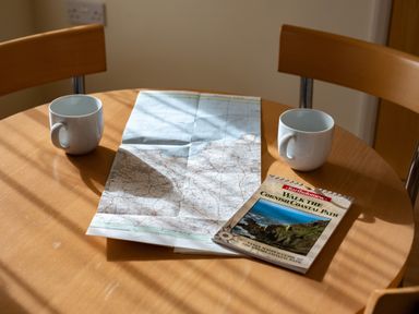 A round wooden table with two white mugs a folded map and a book titled walk the Cornish coastal path at Blue Ridge in Polzeath