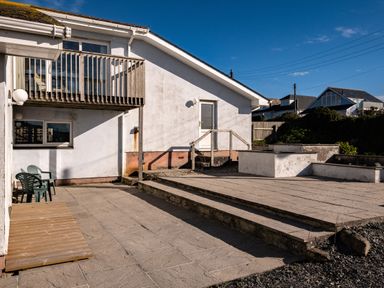 An exterior view of a white building with a wooden balcony plastic chairs on a patio and steps leading to a door at Blue Ridge in Polzeath
