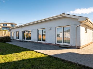 An exterior view of a single-story house with multiple sliding glass doors and a paved walkway surrounded by grass at Blue Ridge in Polzeath