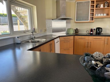 A kitchen with wooden cabinets a black countertop a sink under a window a stove a dishwasher a kettle coffee machine cups and a bowl of decorative items at Blue Ridge in Polzeath