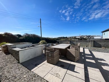 An outdoor patio with a wicker dining table and chairs next to a raised garden bed under a blue sky at Blue Ridge in Polzeath