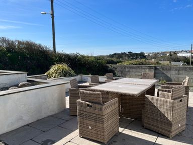 An outdoor patio area with a rectangular table and wicker chairs surrounded by low walls and bushes at Blue Ridge in Polzeath