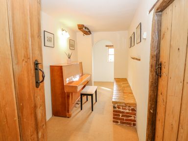 A narrow room with a wooden piano and bench a brick ledge and framed pictures on the walls at Manor Farm House Bacton