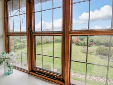 A view of a garden with grass, bushes, and trees through a wooden framed window at Manor Farm House in Bacton
