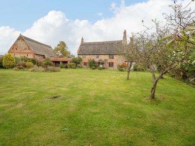A large grass lawn with scattered trees in front of two rustic thatched roof buildings at Manor Farm House in Bacton
