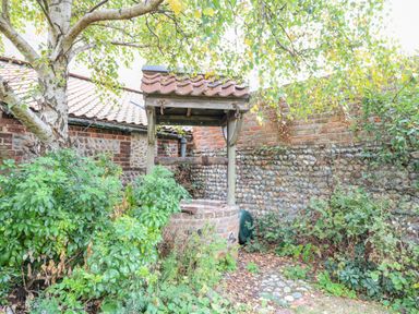 A garden area with a stone and brick well surrounded by plants and a tree next to a stone and brick wall at Manor Farm House in Bacton