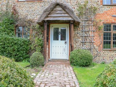 A brick pathway leading to a white wooden door with a wood and thatch porch surrounded by green bushes and a stone and brick wall at Manor Farm House in Bacton