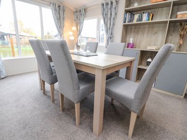 A dining area with six grey upholstered chairs around a wooden table near windows and a shelving unit at Fountain View in Warton near Carnforth