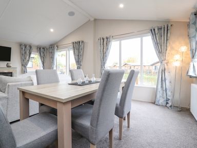 A dining area with a wooden table and six grey upholstered chairs near large windows with patterned curtains at Fountain View in Warton near Carnforth