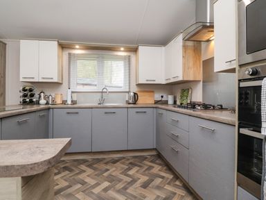 A kitchen with grey lower cabinets and white upper cabinets a window with blinds above a sink and a stove with a vent hood at Fountain View in Warton near Carnforth