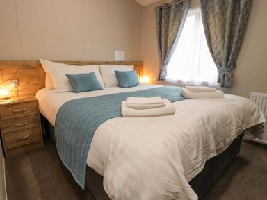 A bedroom with a double bed white bedding blue throw and pillows bedside table with lamp and window with curtains at Fountain View in Warton near Carnforth