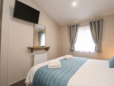 A bedroom with a bed covered by a blue throw and towels on top a wall-mounted TV a mirror above a shelf and a window with grey curtains at Fountain View in Warton near Carnforth